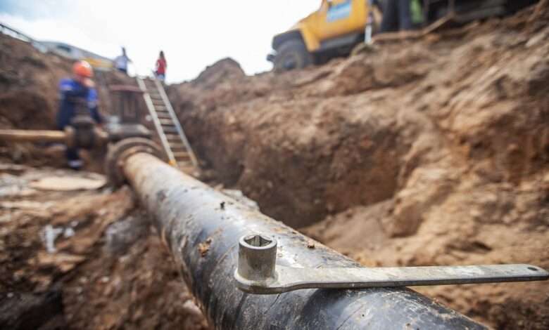 An underground pipeline that's been uncovered. There is a wrench balancing on top of it and some people in the background.