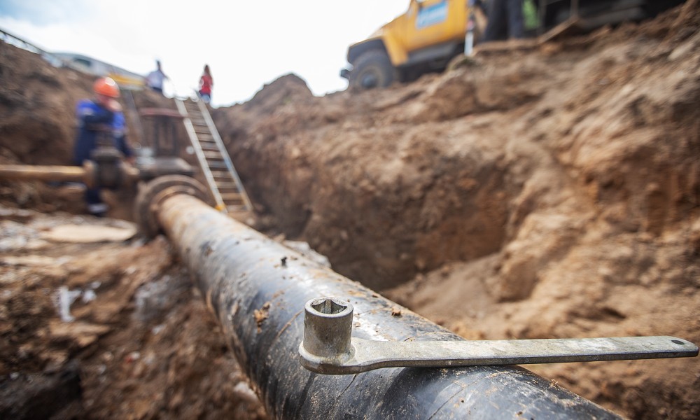 An underground pipeline that's been uncovered. There is a wrench balancing on top of it and some people in the background.