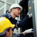 Two employees wearing gloves and hard hats installing industrial equipment in a brightly lit workspace.