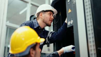 Two employees wearing gloves and hard hats installing industrial equipment in a brightly lit workspace.