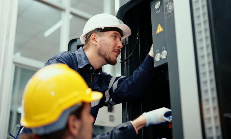 Two employees wearing gloves and hard hats installing industrial equipment in a brightly lit workspace.