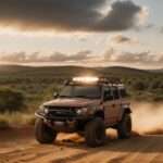 A rugged autonomous off-road vehicle with roof lights kicks up dust on a dirt trail through forested hills at golden hour.