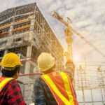 Two contractors wearing safety vests and hard hats look and point up to a high-rise building under construction.