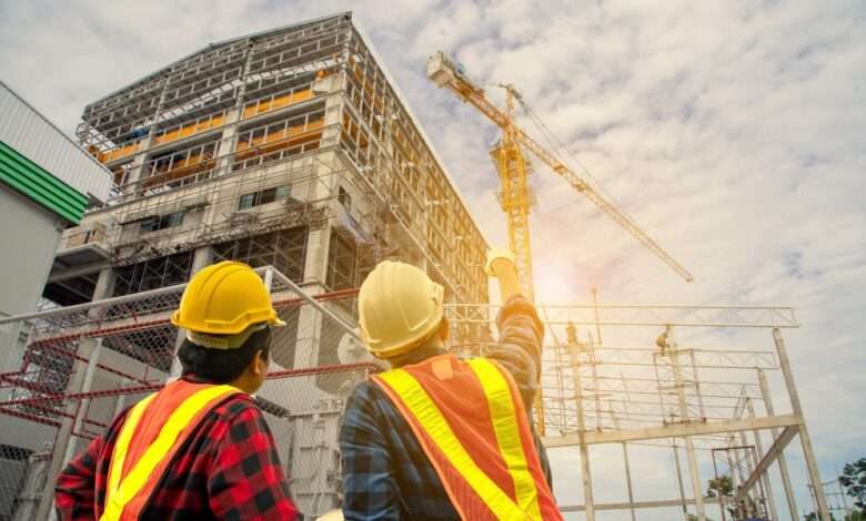 Two contractors wearing safety vests and hard hats look and point up to a high-rise building under construction.