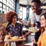 A waiter in an apron serves food to smiling women dining at a restaurant with large windows and brick walls.