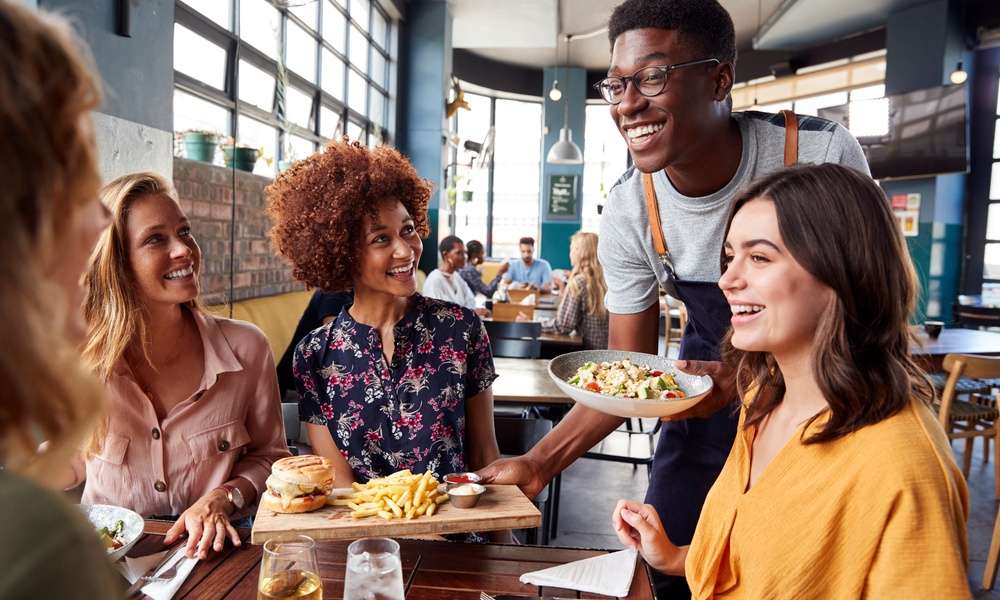 A waiter in an apron serves food to smiling women dining at a restaurant with large windows and brick walls.