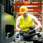 A worker wearing a safety helmet, a yellow vest, and black gloves, holding a wrench next to a toolbox on the floor.