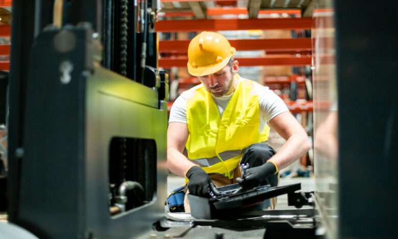 A worker wearing a safety helmet, a yellow vest, and black gloves, holding a wrench next to a toolbox on the floor.