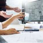 Architects looking at a design for a building on a computer. The desk is covered in building plans and other designs.