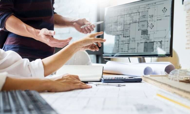 Architects looking at a design for a building on a computer. The desk is covered in building plans and other designs.