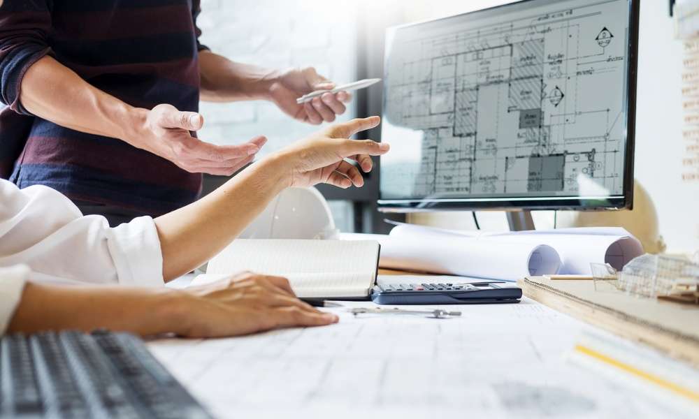 Architects looking at a design for a building on a computer. The desk is covered in building plans and other designs.
