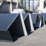 Several pieces of dark gray, angular metal flashing sit in a row on a textured roof awaiting installation.