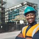 An African American man wearing an orange safety vest and hard hat smiles. Behind him is a building under construction.