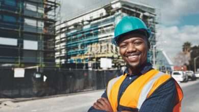 An African American man wearing an orange safety vest and hard hat smiles. Behind him is a building under construction.