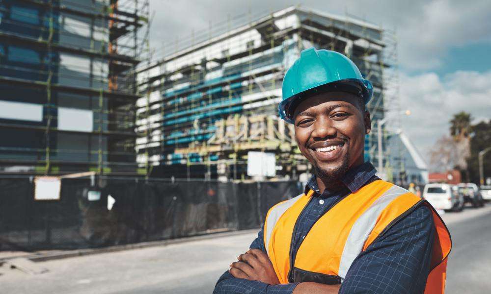 An African American man wearing an orange safety vest and hard hat smiles. Behind him is a building under construction.