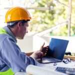 A construction worker wearing a hard hat and a safety vest looks at his laptop while sitting on a project job site.