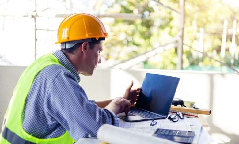 A construction worker wearing a hard hat and a safety vest looks at his laptop while sitting on a project job site.