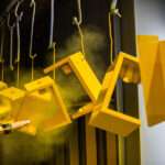 Metal products hang from hooks on a rack. A person uses a powder coating gun to spray the pieces yellow.