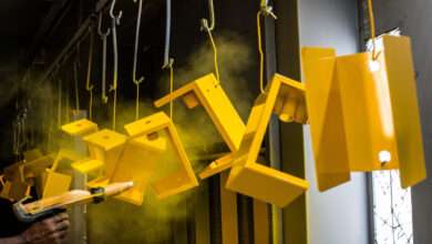 Metal products hang from hooks on a rack. A person uses a powder coating gun to spray the pieces yellow.