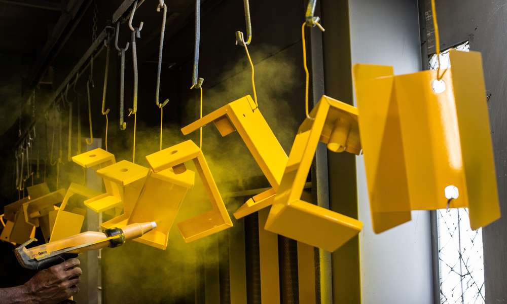 Metal products hang from hooks on a rack. A person uses a powder coating gun to spray the pieces yellow.