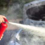 A person uses a red fire extinguisher, discharging white mist near a car with an open hood in an outdoor setting.