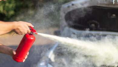A person uses a red fire extinguisher, discharging white mist near a car with an open hood in an outdoor setting.