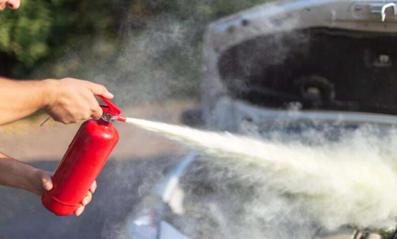 A person uses a red fire extinguisher, discharging white mist near a car with an open hood in an outdoor setting.