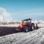 A farmer's field during the winter, with snow on the ground, as a red tractor plows the field into rick, brown dirt.