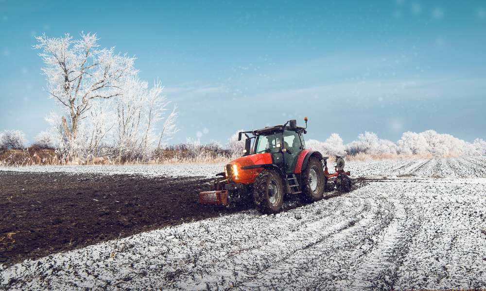 A farmer's field during the winter, with snow on the ground, as a red tractor plows the field into rick, brown dirt.
