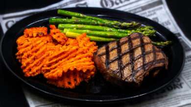 A large black plate with steak, asparagus, and sweet potato fries sits on top of a newspaper.