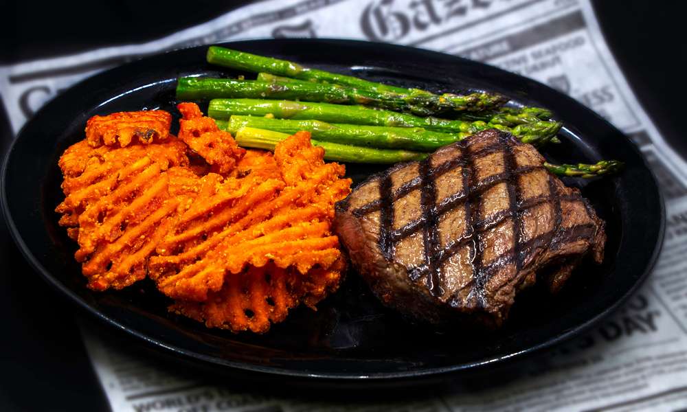 A large black plate with steak, asparagus, and sweet potato fries sits on top of a newspaper.