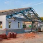 A commercial steel building mid-construction. So far, the structure features gray PBR metal panels, stone siding, and windows.