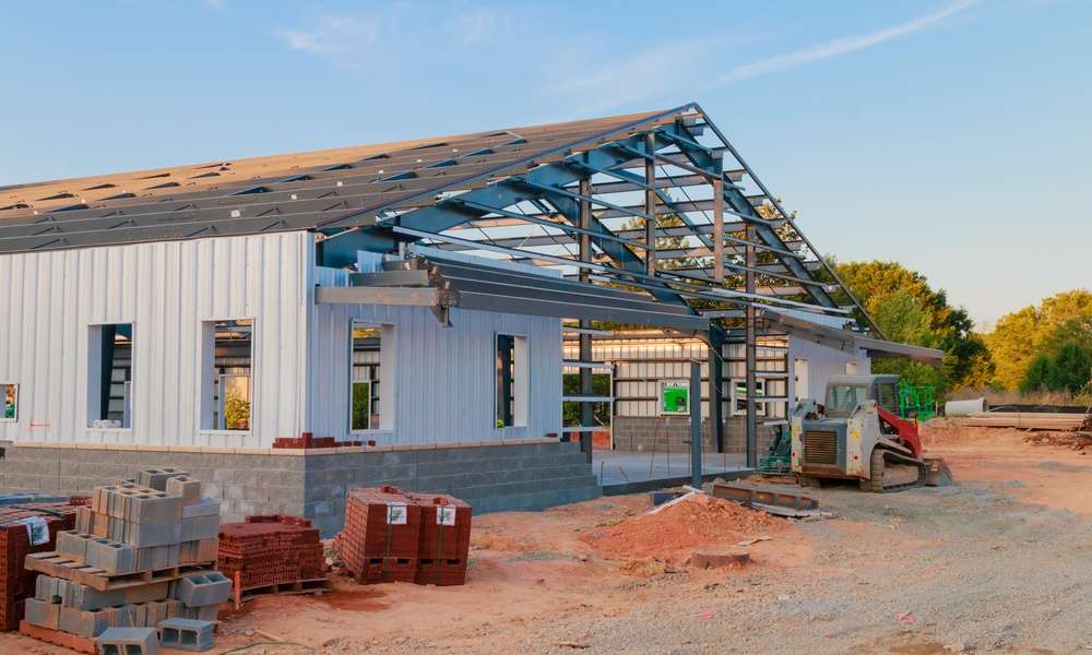 A commercial steel building mid-construction. So far, the structure features gray PBR metal panels, stone siding, and windows.