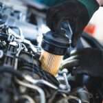 An individual wearing black and green gloves removes a cylindrical oil filter from an open car engine.