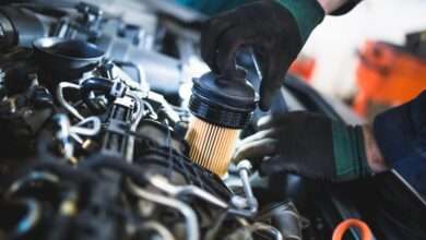 An individual wearing black and green gloves removes a cylindrical oil filter from an open car engine.