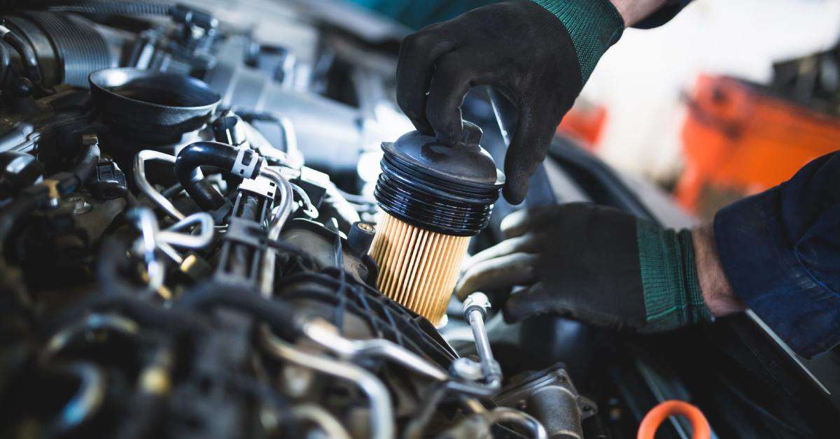 An individual wearing black and green gloves removes a cylindrical oil filter from an open car engine.