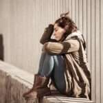 A distressed looking woman is sitting on a concrete barrier; there is a metal fence behind her.