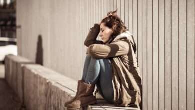 A distressed looking woman is sitting on a concrete barrier; there is a metal fence behind her.