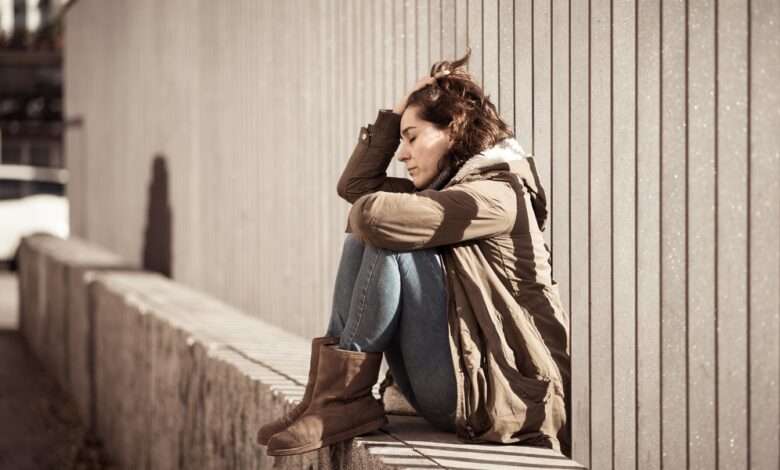 A distressed looking woman is sitting on a concrete barrier; there is a metal fence behind her.