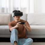 A teenage boy sitting on the floor in front of a couch, wearing a VR headset and holding a controller.