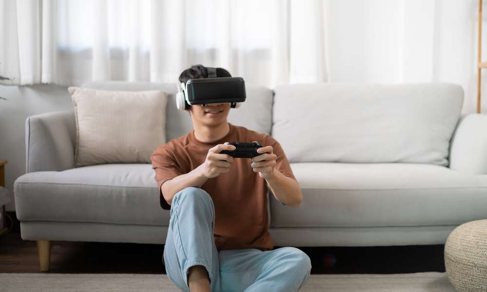 A teenage boy sitting on the floor in front of a couch, wearing a VR headset and holding a controller.