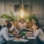 A close-up of a group of young creative business people meeting at the office with a bright yellow light bulb hanging above.