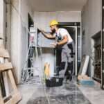 A man with a beard and wearing a yellow hard hat is inspecting an open panel in an unfinished room.