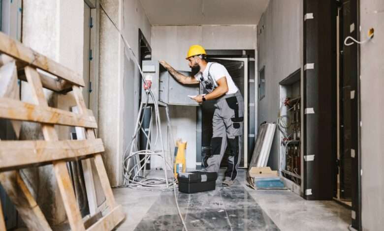 A man with a beard and wearing a yellow hard hat is inspecting an open panel in an unfinished room.