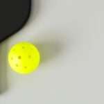 A black pickleball paddle and neon yellow ball on a light gray surface, viewed from above, with subtle shadows.