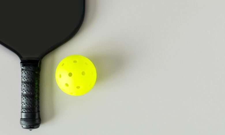 A black pickleball paddle and neon yellow ball on a light gray surface, viewed from above, with subtle shadows.