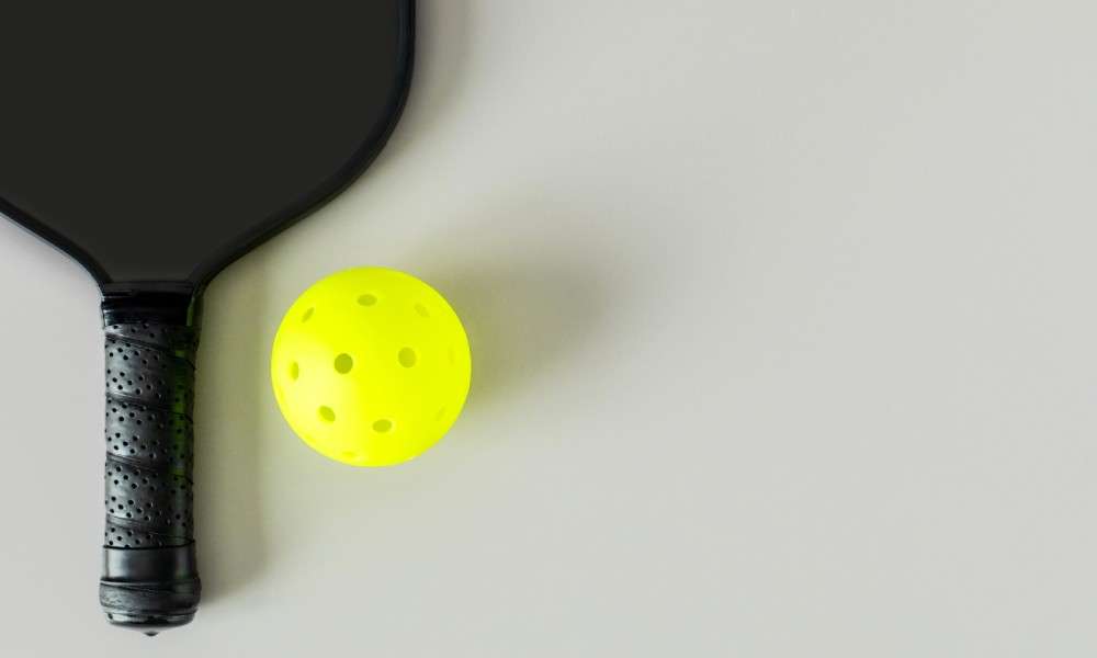A black pickleball paddle and neon yellow ball on a light gray surface, viewed from above, with subtle shadows.