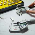 Someone holds a digital micrometer next to a digital caliper. A yellow case holds gauge blocks.