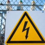 A yellow high-voltage warning sign with a black lightning bolt mounted to a chain-link fence at an electrical substation.
