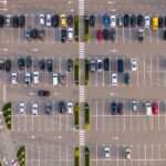 An aerial view of a commercial parking lot. There are many cars parked and they are all of varying colors and designs.